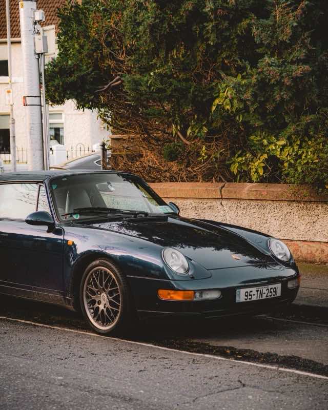 A dark green Porsche 911 993 cabriolet parked at a kerb beside a hedge