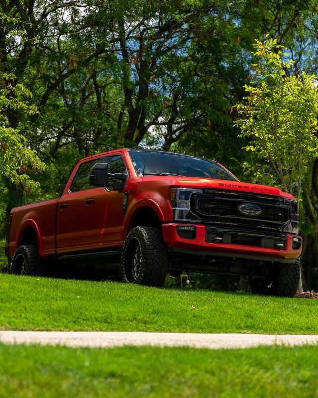 A red Ford Super Duty crew cab pickup on a grass slope under green trees, SUPER DUTY stamped across the leading edge of the hood
