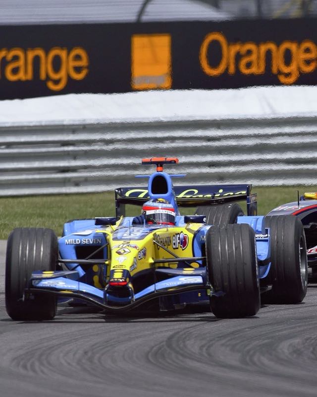A blue and yellow Renault Formula One car cornering ahead of a dark Red Bull at Indianapolis
