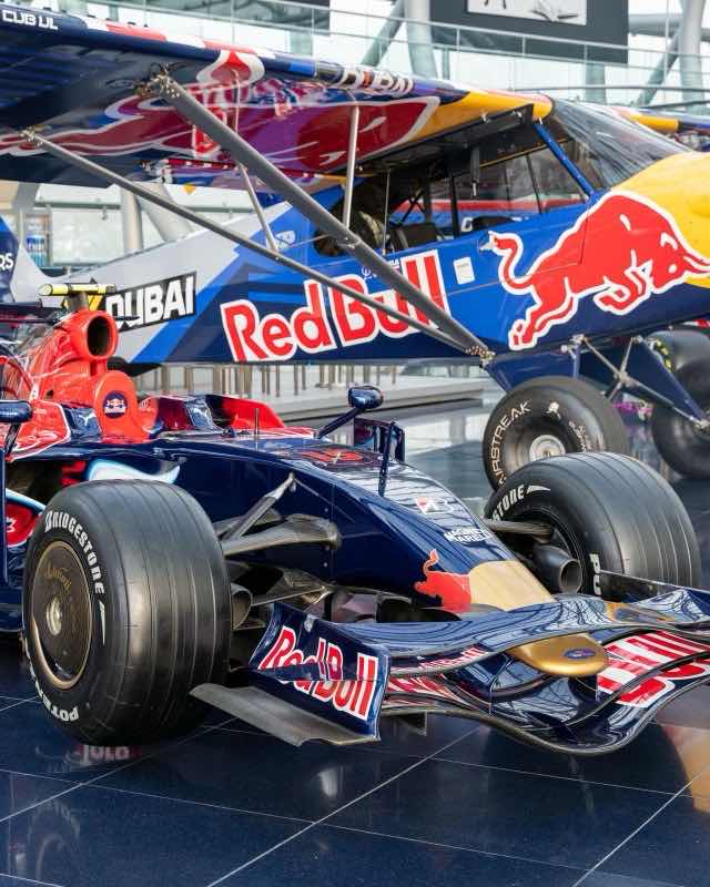 A dark blue and red Toro Rosso Formula One car on display beneath a Red Bull aeroplane in a glass hangar