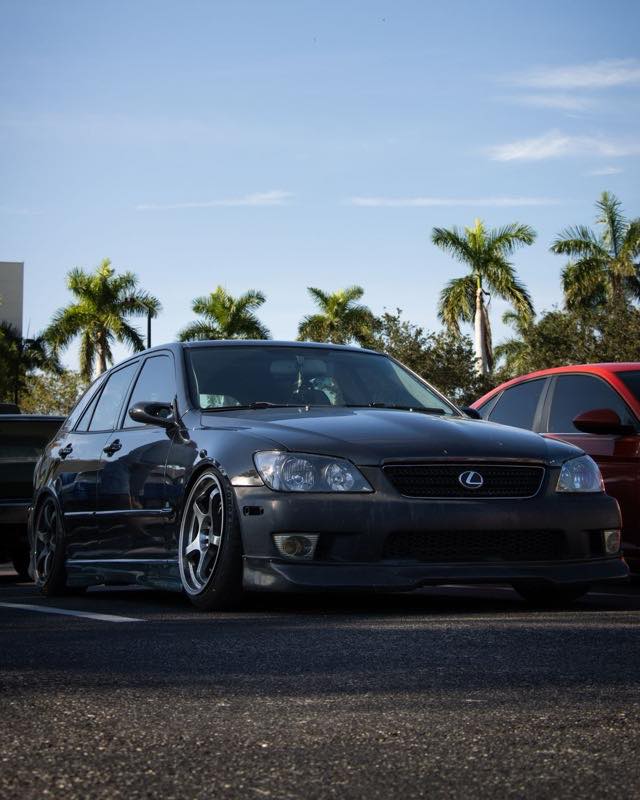 A dark grey first-generation Lexus IS sedan lowered on aftermarket wheels, parked in a palm-lined car park