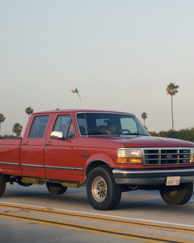 A red Ford crew cab pickup driving along a palm-lined highway at dusk