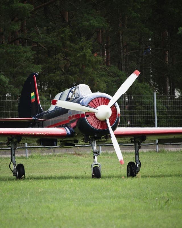A dark blue and red Yakovlev Yak-52 parked on grass, its red engine cowling and two-blade propeller facing the camera