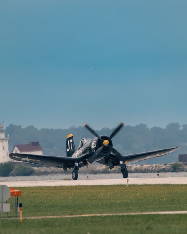 A dark blue F4U Corsair lifting off a runway head on, gull wings bent down at the roots, four-blade propeller turning, a lighthouse behind it