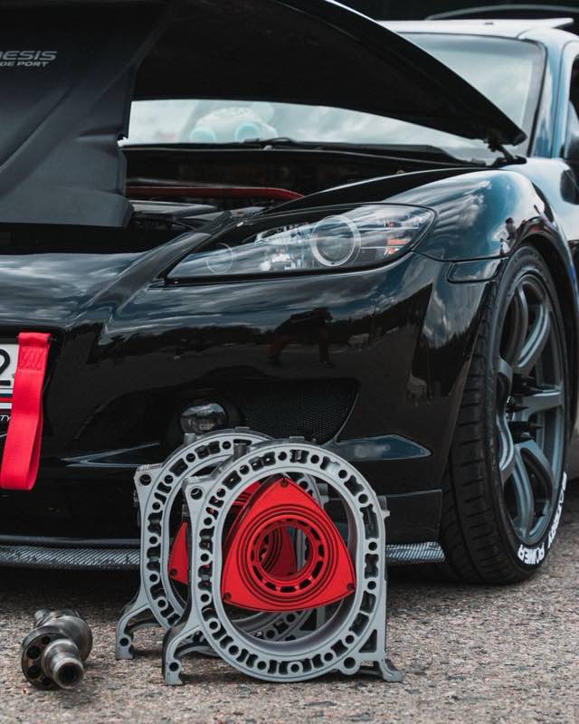 A black Mazda RX-8 with its hood open, a stripped rotor housing and red rotor standing on the ground in front of it