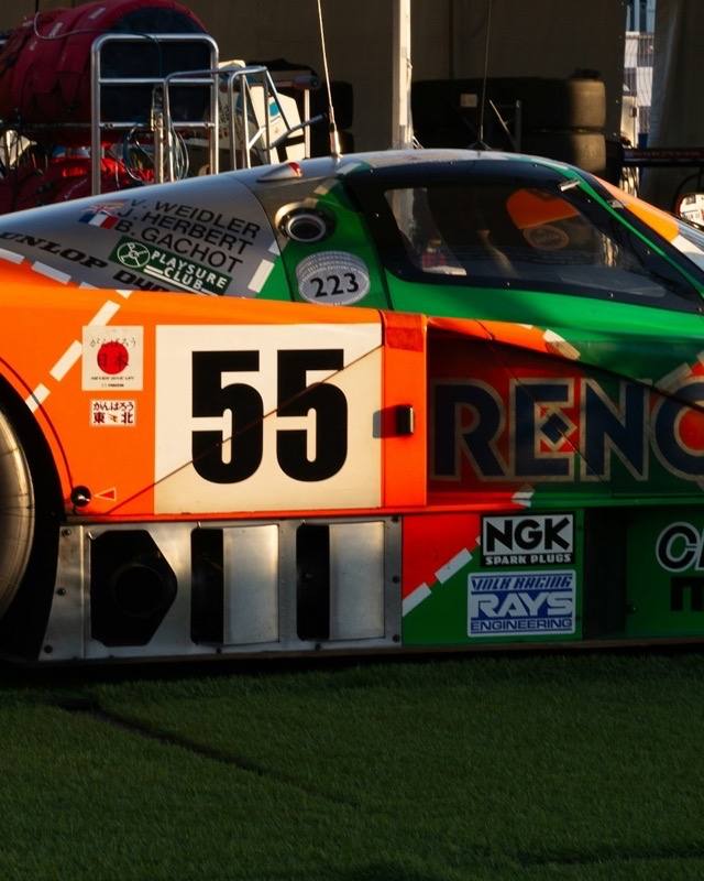 A Mazda 787B in orange and green Renown livery, number 55, parked on grass in a race paddock at sunset