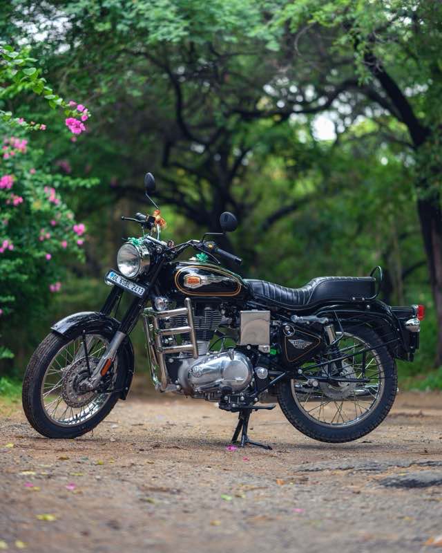 A black Royal Enfield Bullet 350 on its side stand on a tree-lined path