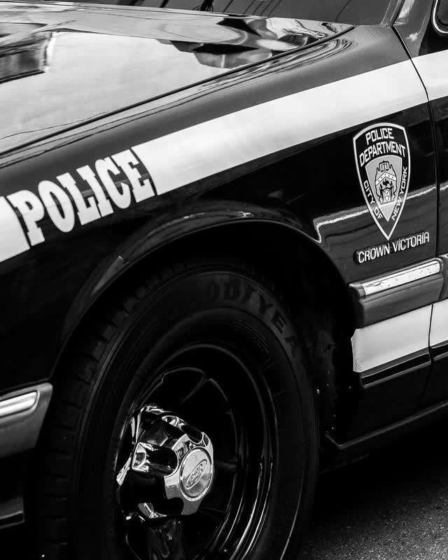 A black and white photograph of a Ford Crown Victoria in NYPD livery, the door badge reading Crown Victoria