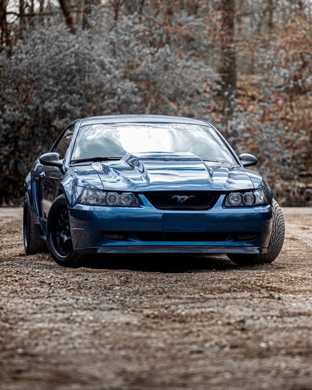 A blue New Edge Ford Mustang seen head on, parked on gravel in front of bare winter woods