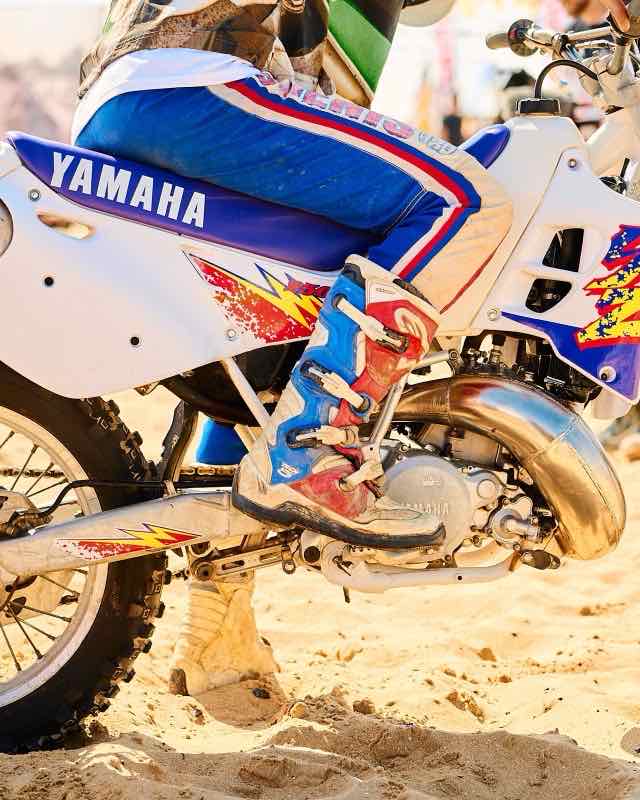 A white and blue Yamaha two-stroke motocross bike on sand, its chrome expansion chamber curving under the engine and a rider's boot on the peg