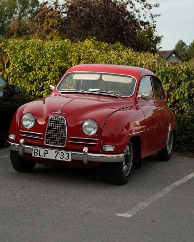 A red two-door Saab 96 with a small oval grille and round headlights, parked on tarmac in front of a hedge