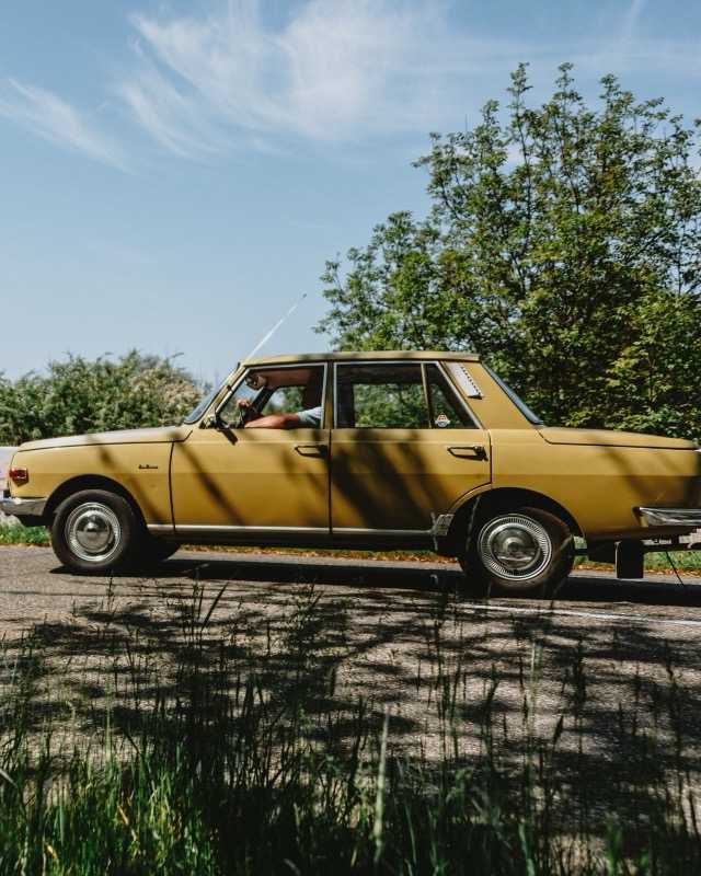 A mustard-yellow four-door Wartburg 353 driving past in profile on a sunlit country road