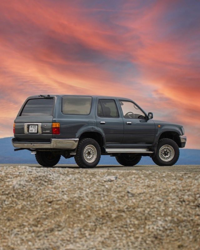 A grey Toyota Hilux Surf parked on gravel under a pink sunset sky