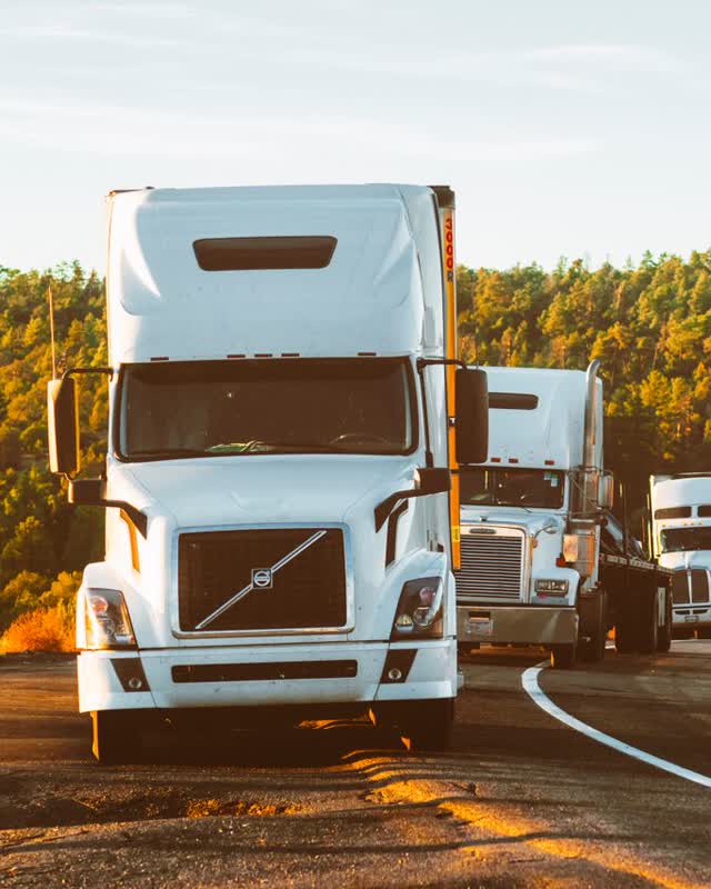 A white Volvo VNL semi-truck leading two other trucks along a forest highway at sunset