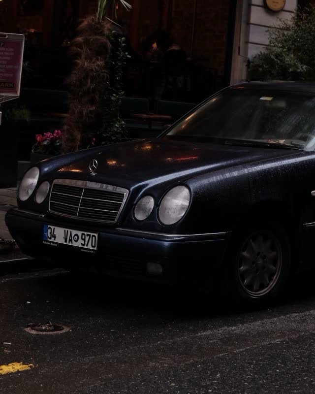 A dark blue Mercedes-Benz E-Class W210 parked on a wet city street at dusk