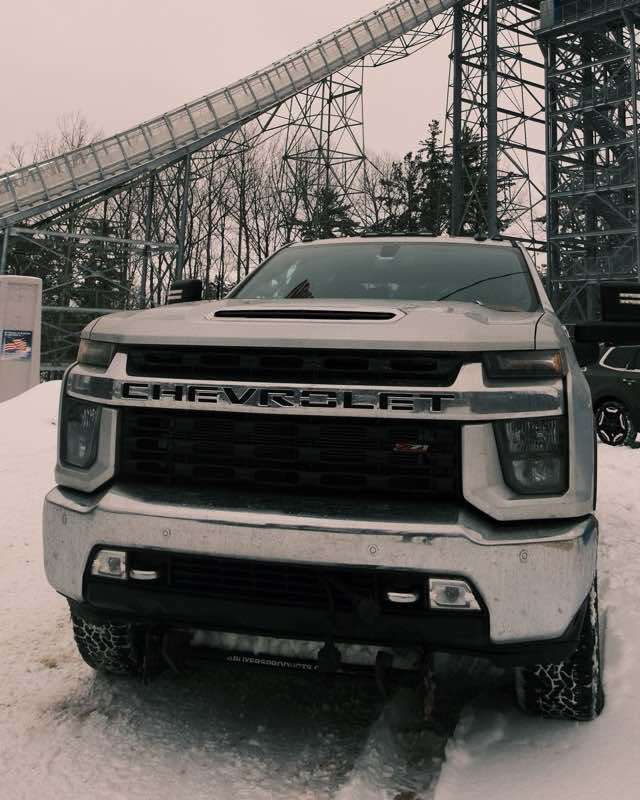 A silver Chevrolet Silverado HD pickup parked in deep snow below a steel ski jump tower