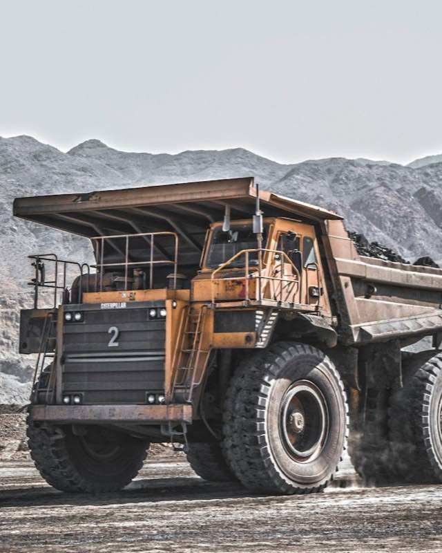 A loaded yellow Caterpillar mining haul truck on a dusty pit road with grey mountains behind
