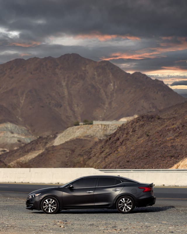 A black Nissan Maxima in profile on a gravel shoulder below a desert mountain at sunset