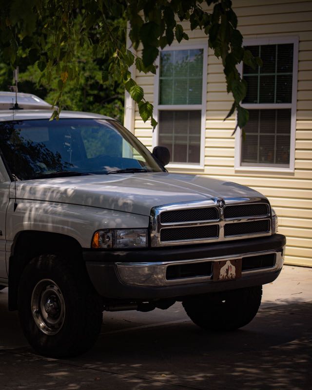 A white second-generation Dodge Ram pickup parked in a shaded driveway beside a yellow house