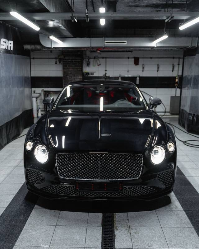 A black Bentley Continental GT facing the camera in a tiled detailing bay with its headlights on