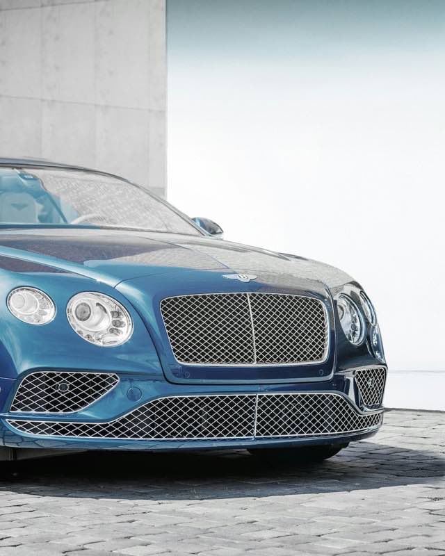 The front end of a blue Bentley Continental GT parked on cobbles beside a concrete wall
