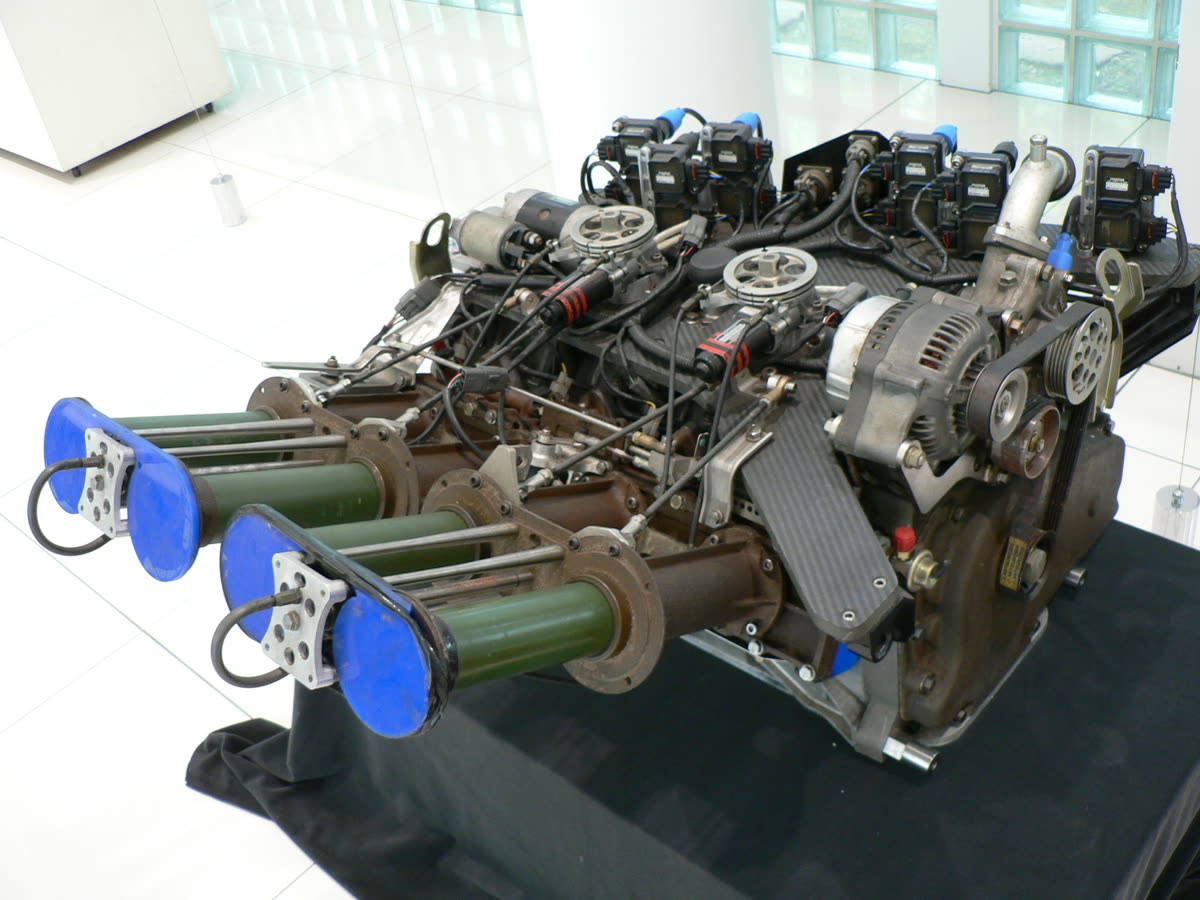 Photograph of a Mazda R26B four-rotor racing engine on display, showing four telescoping variable-length intake trumpets with blue caps on the left, banks of ignition coils along the top, and the alternator and belt drive on the right