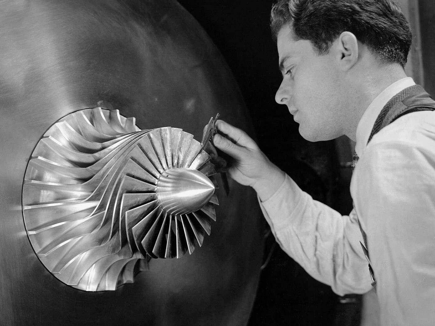 Black and white 1944 photograph of a young NACA researcher in shirtsleeves measuring the blade thickness of a polished centrifugal supercharger impeller with a caliper, the curved impeller vanes clearly visible