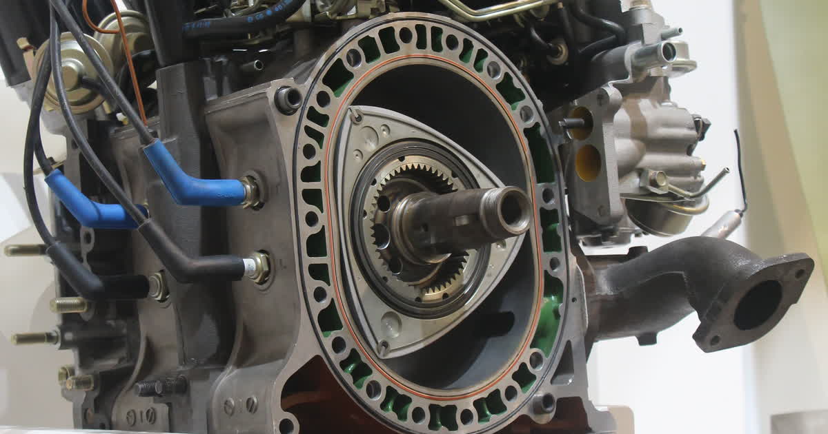 Sectioned Mazda 13B rotary engine on museum display, cut through one rotor housing with the rotor removed, showing the empty two-lobe epitrochoid bore, its orange sealing bead, green coolant passages in the housing wall, and the eccentric shaft passing through the fixed stationary gear on the side housing