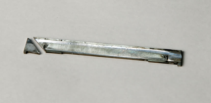Photograph of a Mazda 13B apex seal: a thin narrow metal strip roughly the width of the rotor, lying next to the small triangular key that locates it, on a plain background